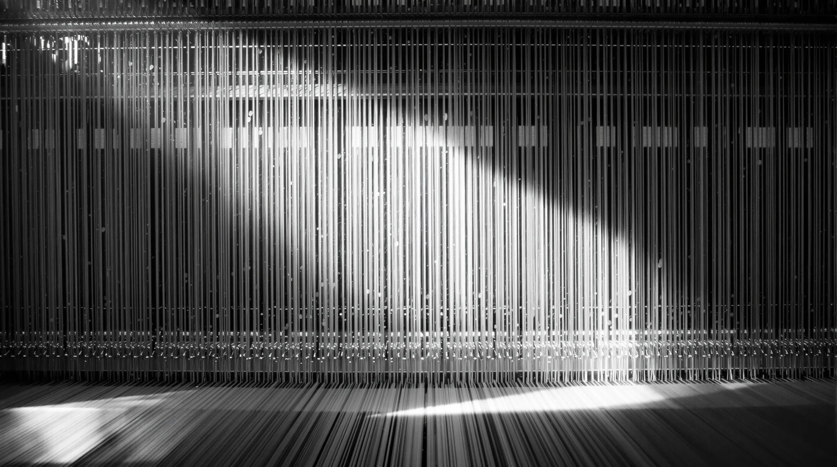 A vintage Jacquard loom showing dense vertical warp threads under tension, photographed in dramatic black and white. Metaphor for how a language model weaves text token by token across many parameter threads.